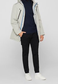 Parka imbottita grigia chiara con cappuccio, dotata di dettagli neri sulle zip. Abbinata a pantaloni cargo neri e sneakers bianche. Design minimalista.