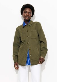 Femme aux cheveux courts et bouclés portant une veste olive à boutons sur une chemise à rayures bleues et un pantalon blanc, se tenant devant un fond blanc.