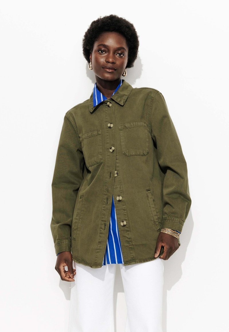 Femme aux cheveux courts et bouclés portant une veste olive à boutons sur une chemise à rayures bleues et un pantalon blanc, se tenant devant un fond blanc.
