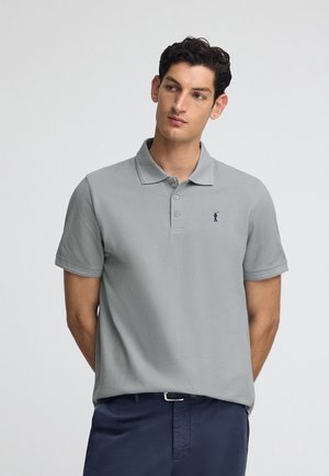 Polo shirt gris clair avec un col, trois boutons et un petit logo bleu marine. Tissu lisse à manches courtes, associé à un pantalon sombre.