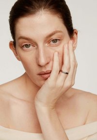 Anillo de plata con un acento negro, llevado en un dedo, combinado con una blusa de tono neutro y con los hombros al descubierto; la mano del modelo descansa sobre su rostro.