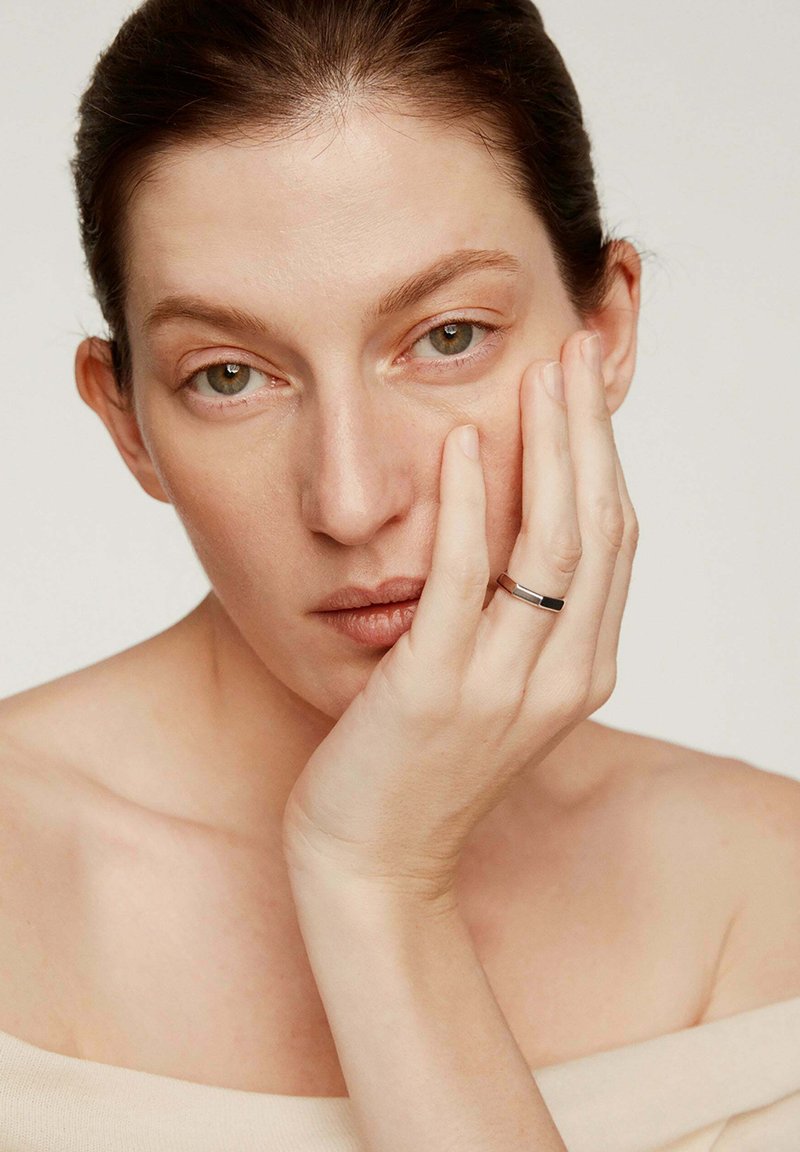 Anillo de plata con un acento negro, llevado en un dedo, combinado con una blusa de tono neutro y con los hombros al descubierto; la mano del modelo descansa sobre su rostro.