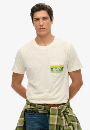Cream short-sleeve t-shirt with a green and yellow pocket logo; soft texture, crew neckline, paired with a green plaid shirt tied at the waist.