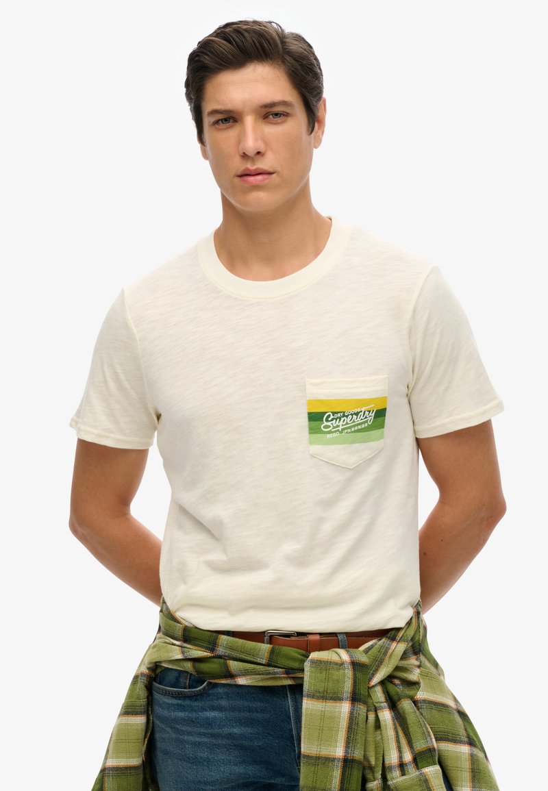 T-shirt crema a maniche corte con logo a tasca verde e giallo; texture morbida, scollatura a girocollo, abbinato a una camicia a quadri verde legata in vita.