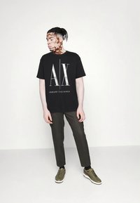 Μαύρο βαμβακερό T-shirt με ασημί λογότυπο "AX" και κείμενο "ARMANI EXCHANGE", συνδυασμένο με σκούρο πράσινο παντελόνι στενής γραμμής και ελιά παπούτσια.