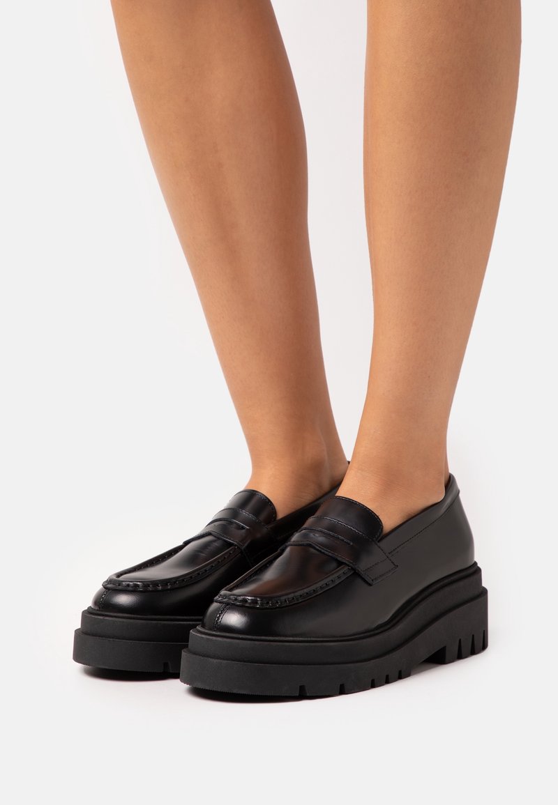 Jambes portant des mocassins à plateforme en cuir noir avec des semelles en caoutchouc épaisses et rainurées, posées sur un fond blanc.