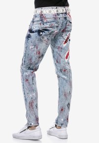 Verbleekte blauwe denim jeans met verfsplatselpatronen in rood en zwart. Afgewerkt met een witte studded riem en accentueert met witte sneakers.