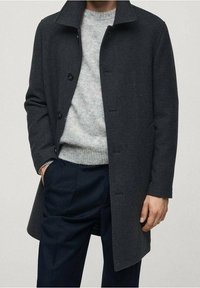 Abrigo de lana gris oscuro con cuello, botones frontales y bolsillos laterales, llevado sobre un suéter gris claro y pantalones oscuros.