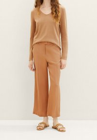 Beiger Strickpullover mit V-Ausschnitt, kombiniert mit weiten, beige Hosen und braunen Sandalen. Die Materialien wirken weich und leicht.