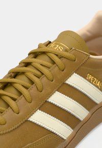 Καφέ δερμάτινο αθλητικό παπούτσι με λευκές ρίγες, χρυσό κείμενο "SPEZIAL" και καφέ κορδόνια που ταιριάζουν, σε λευκό φόντο.