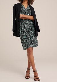 Zwarte blazer over een groene bloemenjurk met een getailleerde taille. Hakken met een zwarte enkelband maken de outfit compleet.