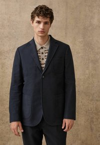 Marineblauer Leinenblazer mit einem einzelnen Knopf, Reverskragen und zwei vorderen Taschen, getragen über einem hellbraunen gemusterten Hemd. Texturierter Hintergrund.