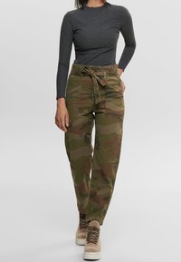Frau in grauem Langarmshirt, camouflierte, hochtaillierte Hose mit gebundenem Gürtel und beigen Schnürstiefeln, die vor einem grauen Hintergrund geht.