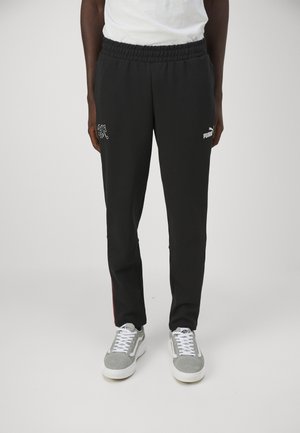Schwarze Jogginghose mit elastischem Bund, ausgestattet mit einem weißen gestickten Logo und roten Seitenstreifen. Getragen mit grauen Sneakers.
