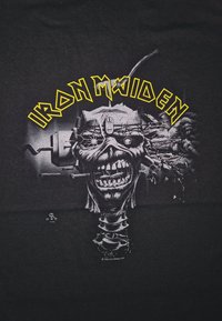 Camiseta negra con un gran gráfico de un esqueleto con un diseño mecánico. "IRON MAIDEN" en letras amarillas en negrita encima de la imagen.