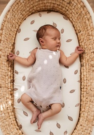 Ein helllila gestrickter Baby-Overall mit Knopfakzenten, der auf einer gepolsterten Oberfläche mit braunen Blattmustern liegt, in einem geflochtenen Korb.