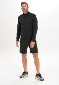 Svart zip-up träningsjacka med hög krage och fickor, kombinerad med matchande shorts. Materialet verkar lätt och flexibelt.
