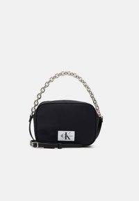 Bolso rectangular negro hecho de nylon, con un asa de cadena, correa ajustable y una parche de marca que presenta las iniciales "CK."