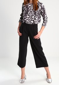 Pantaloni neri a gamba larga abbinati a una blusa a maniche lunghe grigia e bianca con motivo a fantasia e dettagli in raso; tacchi argentati lucidi completano il look.