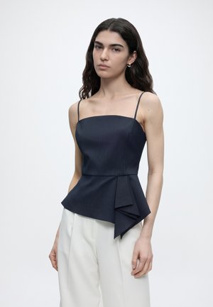 Nő hosszú, hullámos sötét hajjal, tengerészkék vékony csíkos ujjatlan topot viselve, aszimmetrikus fodorral és fehér, széles szárú nadrággal.