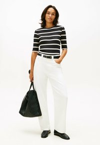 Top de rayas en blanco y negro con canalé, pantalones anchos blancos, cinturón negro y bolso de tote negro texturizado. Modelo con zapatos negros, manos en los bolsillos.