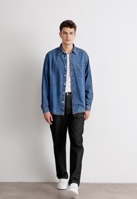 Camicia in denim azzurra con bottoni, con dettaglio della tasca, abbinata a pantaloni scuri e sneaker bianche. Sfondo semplice, vista a corpo intero.