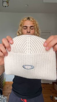 Berretto lavorato a maglia in beige chiaro, con una texture a coste verticali e un risvolto, arricchito da un sottile logo grigio nella parte frontale.