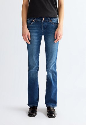 Személy közepesen koptatott bootcut farmert visel réz gombokkal és fekete mokaszinnal, egyszerű világos háttér előtt áll.