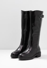 Bottes noires montant jusqu'au genou avec une finition brillante, présentant un panneau texturé à l'arrière, une fermeture éclair sur le côté et une semelle en caoutchouc épaisse.