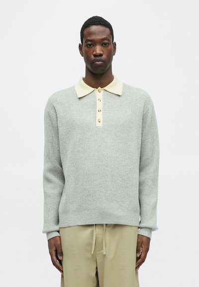 Pull en tricot gris avec un col crème et quatre boutons beiges. Il possède des manches longues et des poignet en côtes, associé à un pantalon beige.
