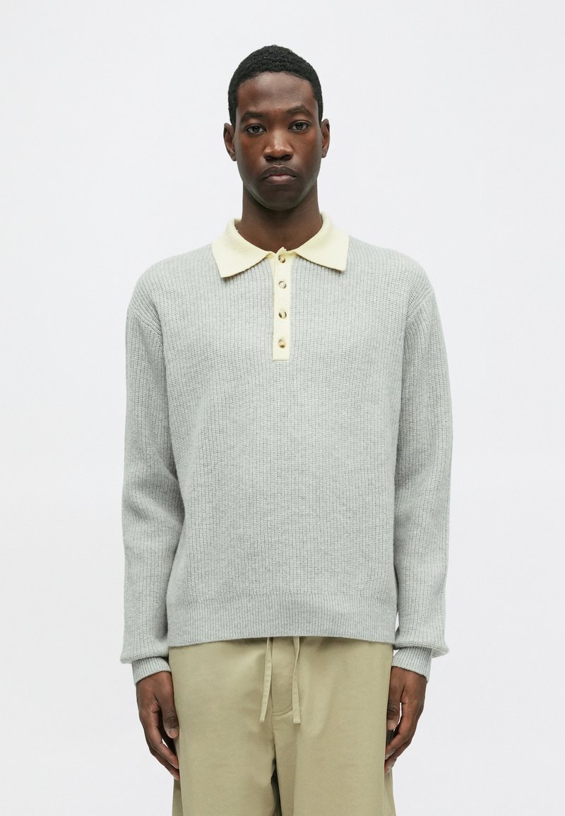 Hallid hallist lõngast kootud kampsun, millel on kreemivärvi krae ja neli beeži nuppu. Omab pikki varrukaid ja soonikuid, koos beežide pükstega.