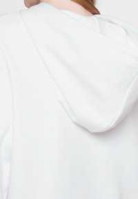 Gros plan d'une personne portant un sweat à capuche blanc uni, montrant le dos et la capuche avec des cheveux longs partiellement visibles.