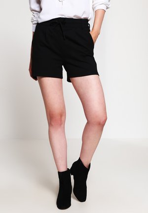 Pantaloni corti neri realizzati in tessuto morbido con una vita regolabile, caratterizzati da una vestibilità sartoriale. Indossati con stivali neri alla caviglia e una camicia bianca.