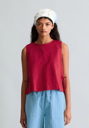 Femme portant un haut rouge sans manches avec des liens sur les côtés, un pantalon rayé bleu et blanc, et un foulard à fleurs blanc, debout devant un fond uni.