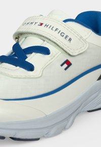 Baskets blanches avec lacets et doublure bleus, bride en velcro étiquetée Tommy Hilfiger, et petit logo drapeau Tommy Hilfiger sur le côté.