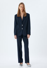 Navy tailored blazer met grote zilveren knopen en uitgesneden revers, gecombineerd met bijpassende broek. Gladde stof met een gestructureerde pasvorm.