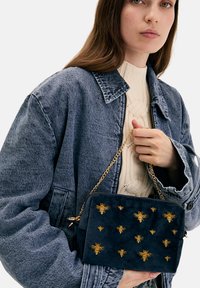 Mujer vestida con chaqueta de mezclilla azul y suéter crema sosteniendo un bolso azul marino con bordado de abeja dorada y cadena como correa.