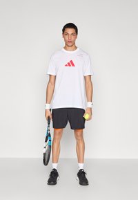 Valge sportlik särk punase Adidas logo ja mustad lühikesed püksid oranži äärega, mustad jalanõud, tennisereket ja kollane tennispall käes.
