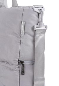 Bolsa gris acolchada con un acabado texturizado, que presenta un cierre de cremallera plateado, una etiqueta de marca y una correa ajustable resistente.