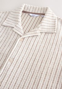 Camicia con bottoni in tessuto strutturato con base color crema e sottili strisce marroni. Dettagli notevoli includono un colletto alla francese e bottoni beige.