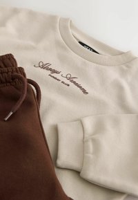 Beige sweatshirt with ribbed cuffs and "Always Awesome Sunday Club" text paired with brown pants with elastic waistband and drawstring.