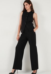 Vesta fără mâneci neagră cu cinci nasturi și buzunare, asociată cu pantaloni negri cu picioare largi. Tessă netedă, design ajustat și stil minimalistic.