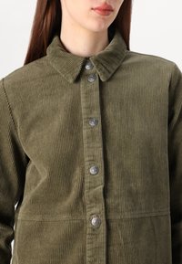 Camisa-jacket de ganga em verde oliva, com fecho de botões à frente, colarinho pontudo e nervuras verticais texturizadas. Apresenta botões prateados e um corte ajustado.