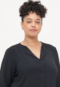 Mustapaita, jossa on V-aukko ja pitkät hihat, hienovaraisella tekstuurilla ja rento istuvuus, korvissa nappikorvakorut.