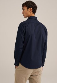 Mornarsko modra srajca z gumbi in dolgimi rokavi, prilagojena kroja, z enim zadnjim delom in subtilno teksturo, v kombinaciji s peščenimi bež hlačami.