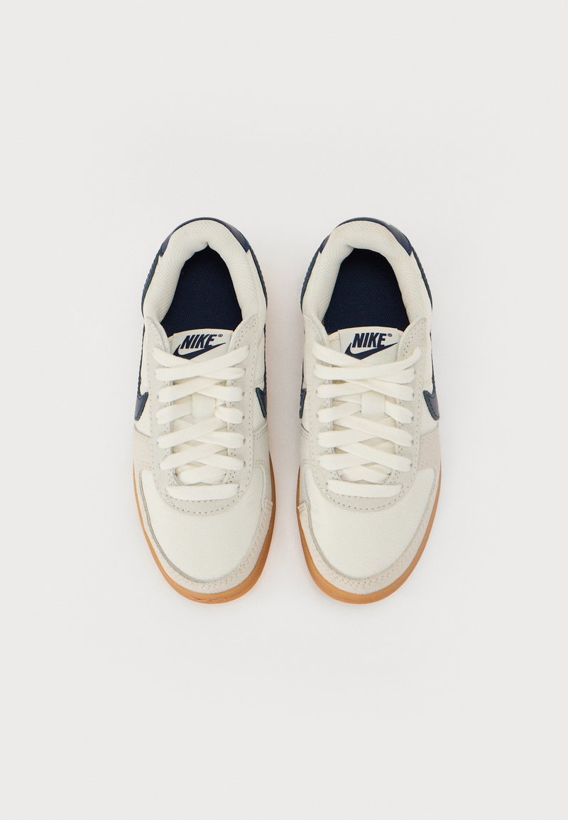 Zapatillas Nike con un upper de lona color crema, acentos en azul marino y suela de goma. Presentan cordones blancos y un logo tejido en la lengüeta.