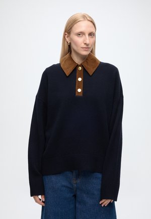 Femme blonde portant un pull bleu marine avec un col en velours côtelé marron et des boutons dorés, associé à un jean bleu, debout devant un fond uni.