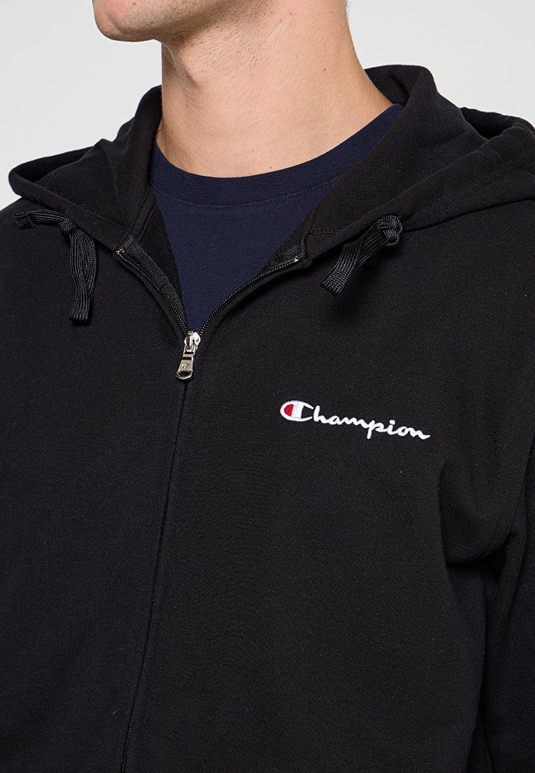 Czarna bluza z kapturem zapinana na zamek, wykonana z miękkiego materiału, z kapturem ze ściągaczem, z białym haftowanym logo "Champion" po lewej stronie klatki piersiowej.
