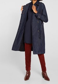 Denim trench coat v tmavě modré barvě, s knoflíkovým zapínáním, nastavitelným páskem v pase a dlouhými rukávy. Nosený přes tmavou šaty a červené semišové kalhoty.