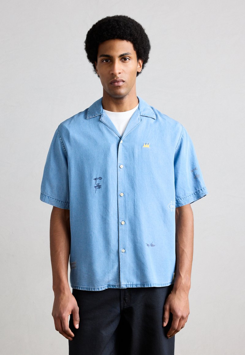 Άνδρας που φοράει ανοιχτό μπλε πουκάμισο με κοντά μανίκια, κουμπωτό μπροστά, με μικρά αφηρημένα σχέδια προσώπου και κορώνας, σε συνδυασμό με μαύρο παντελόνι, στέκεται μπροστά σε απλό φόντο.